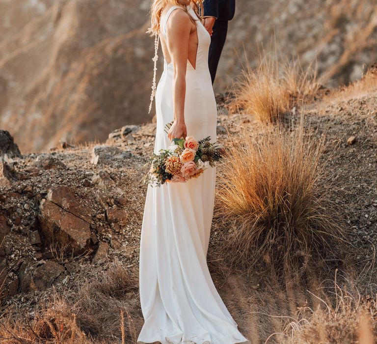 Groom leads bride along the cliffside as she looks back at the camera