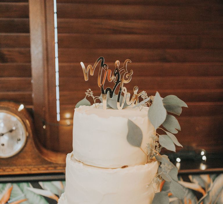 A three tiered white buttercream wedding cake has a topper reading mrs & mrs and is decorated with foliage at Crab & Lobster fairytale wedding.