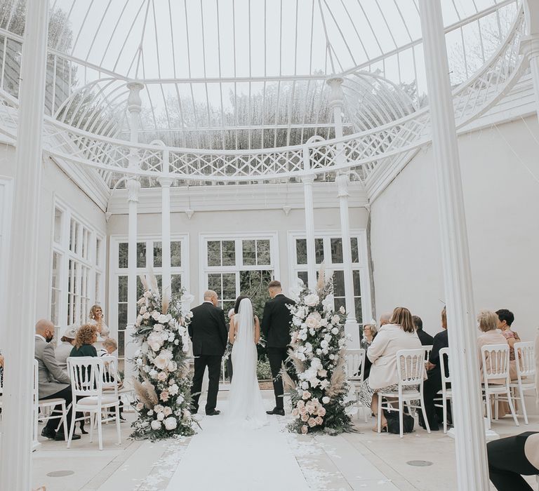 Bride and groom stand at end of white carpeted aisle in the glass conservatory for wedding ceremony at Came House Dorset