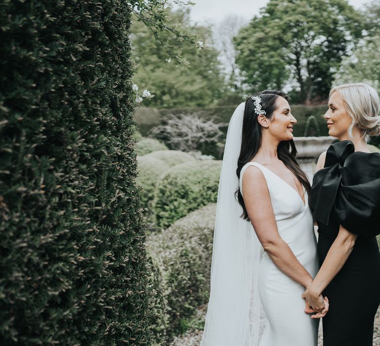 Bride with long dark hair held back with silver hair accessory wearing white Pronovias wedding gown and veil stands in grounds of Came House Dorset holding hands with maid of honour in black one-shoulder gown