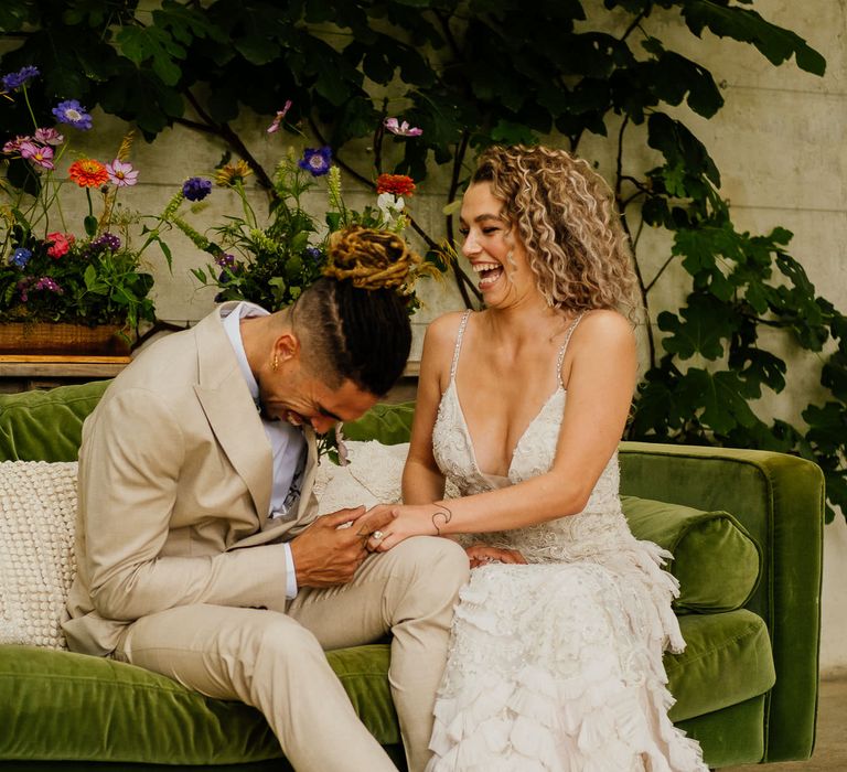 Groom in a beige suit and bride in an embellished wedding dress with straps and ruffles laughing on a green velvet sofa 