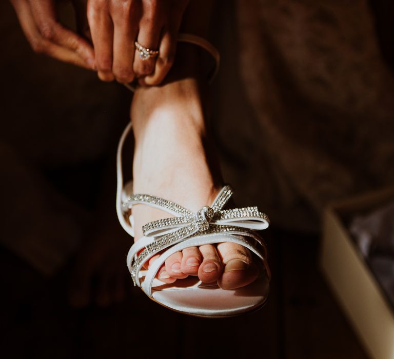 Bride wears pearl bracelet whilst doing up strappy bridal shoes with diamanté encrusted bow to the front  