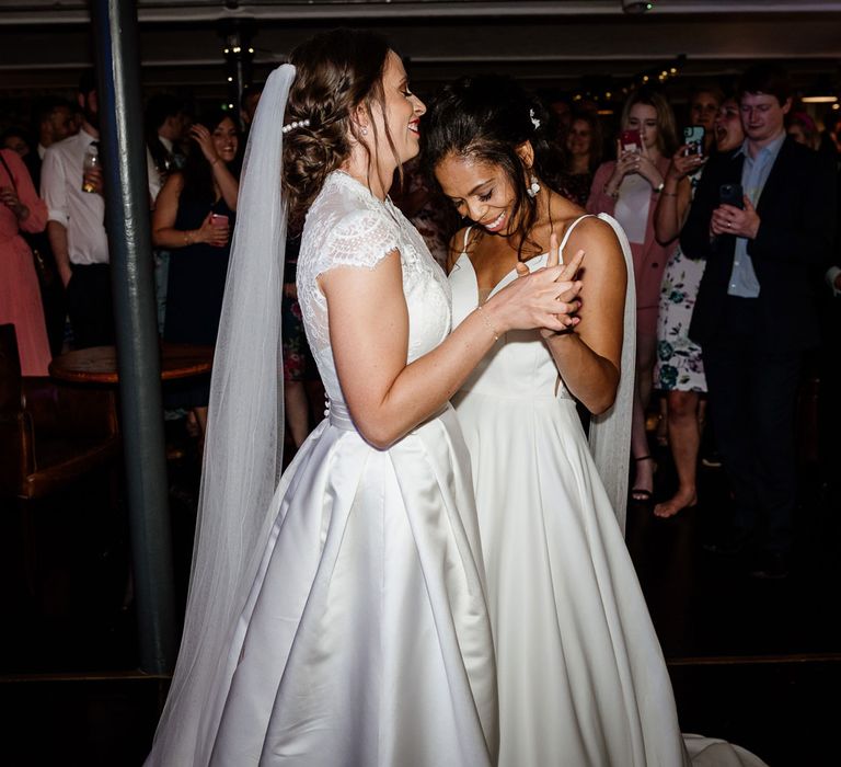 Smiling bride in white cami wedding dress with shoulder veil details dances with bride in white lace top capped sleeve wedding dress an veil during wedding reception at The West Mill Derby