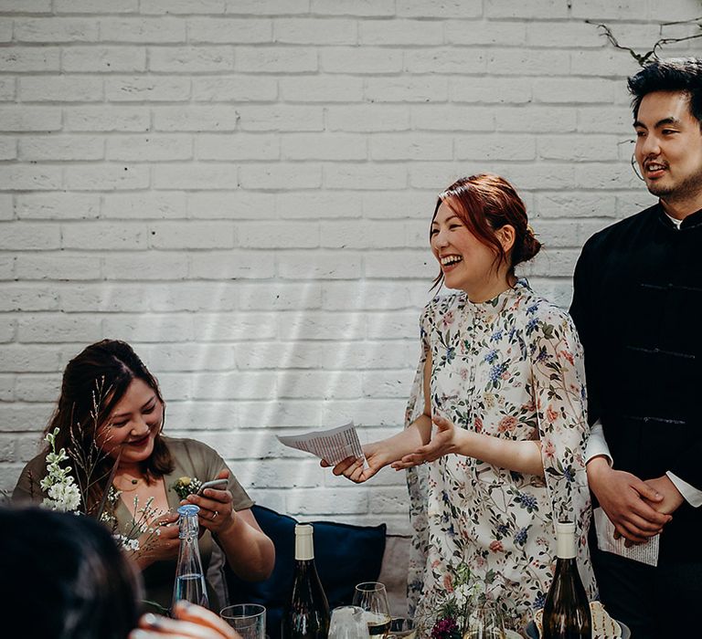 East Asian bride in a floral wedding dress and groom in a navy suit jacket delivering their wedding speeches together 