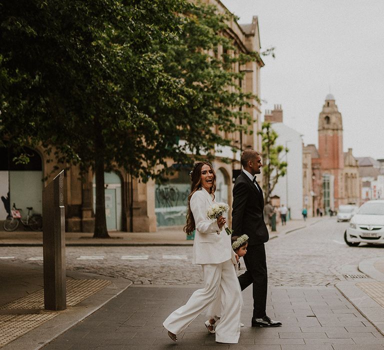 Bride in a wide leg trousers and blazer suit walking through Sheffield with her flower girl daughter and groom in a tuxedo 