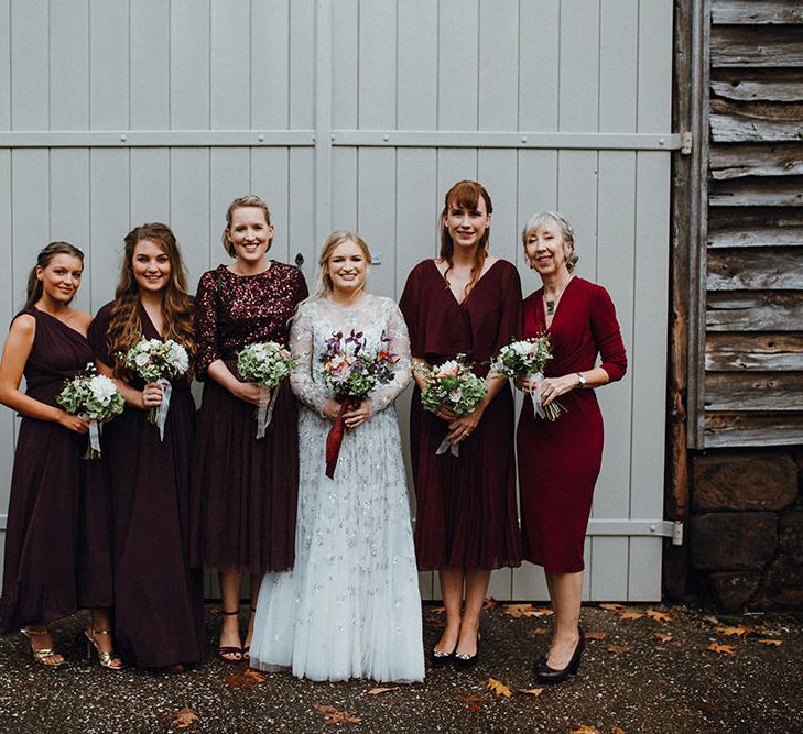 Bride in a tulle wedding dress with silver embroidery standing with her bridesmaids in different style, fabric and length burgundy dresses