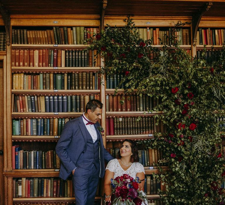 Portrait of a groom in blue three-piece suit with burgundy tie looking at his bride in a wheelchair wearing lace bridal separates in the library at Dulwich College 