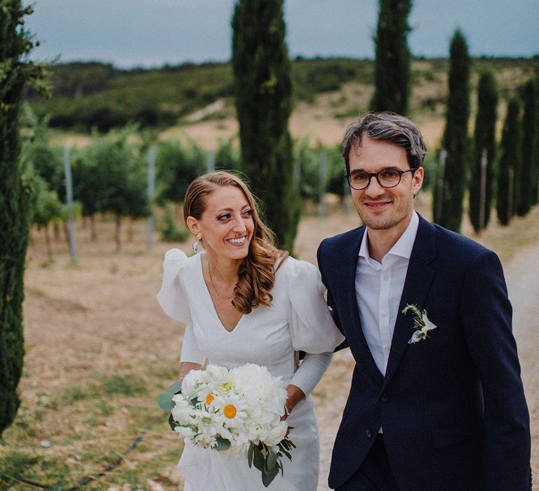 Bride & groom laugh as they walk through golden fields as groom wears glasses 