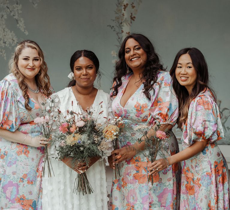 Bride in white long mesh sleeve spotted wedding dress stands with bridesmaids in multicoloured floral dresses all holding mixed bouquets outside Victorian summerhouse at Wasing Park wedding