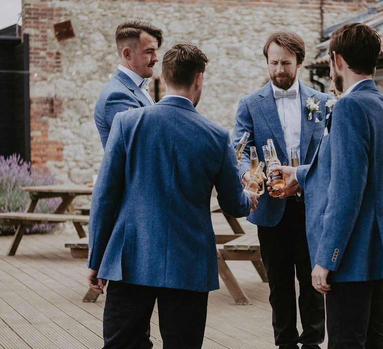 Groomsmen in blue suit jackets and bow ties all clink bottles of Corona before Isle of Wight wedding with macrame wedding decor