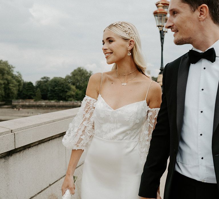 Portrait of a groom in tuxedo with white buttonhole flower holding hands with his bride in a satin and lace wedding dress on Putney Bridge 