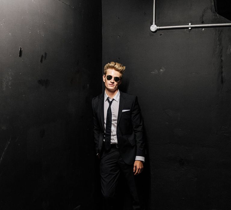 Groom wears black suit, white shirt and black tie with ray ban sunglasses 