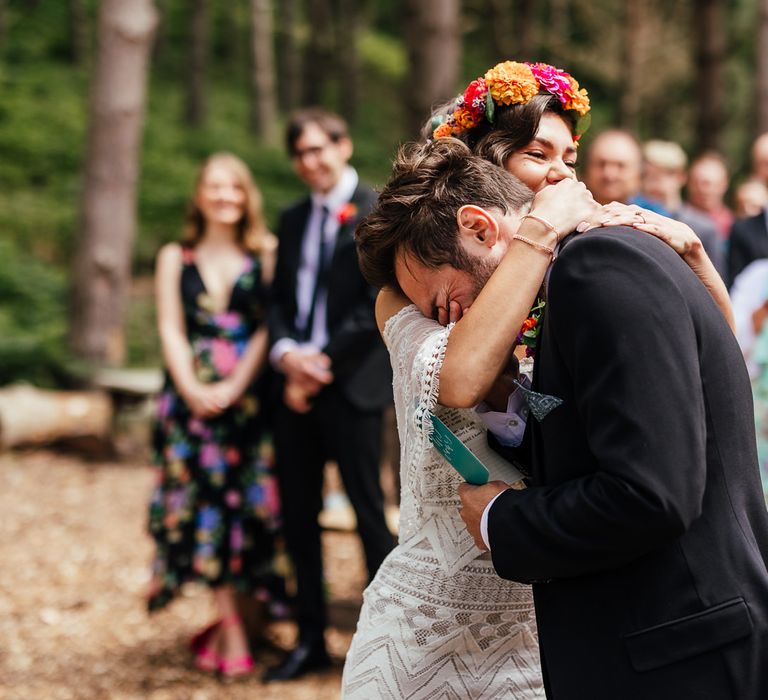 Bride in off the shoulder Grace Loves Lace wedding dress and flower crown hugs groom and he holds vows during woodland wedding ceremony