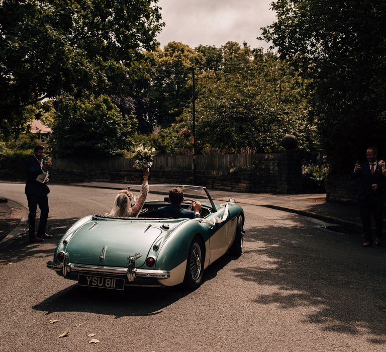 Bride holds up bouquet in the air and groom drives them both away from the church in vintage blue and white sports car after wedding ceremony