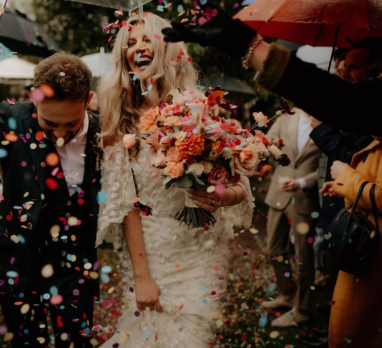 Bride in floral applique wedding dress holding peachy bridal bouquet smiles whilst walking with groom in dark suit carrying umbrella as guests throw multicoloured confetti 