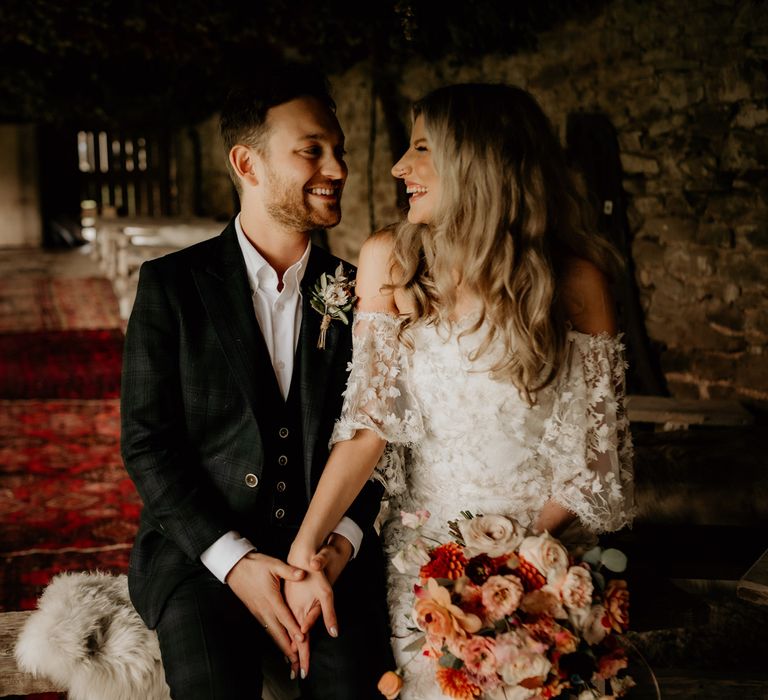 Laughing bride in bardot wedding dress holding mixed autumnal bridal bouquet sits holding hands with groom in dark three piece suit with floral buttonhole in rustic wedding ceremony room