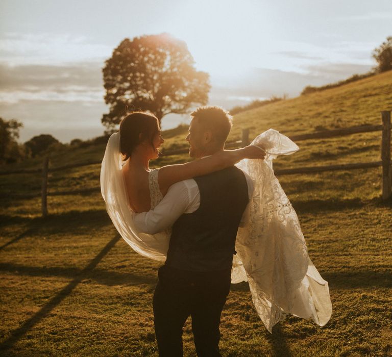 Groom in white shirt and waistcoat holds bride in lace wedding dress and veil during golden hour at Hope Farm in Dorset