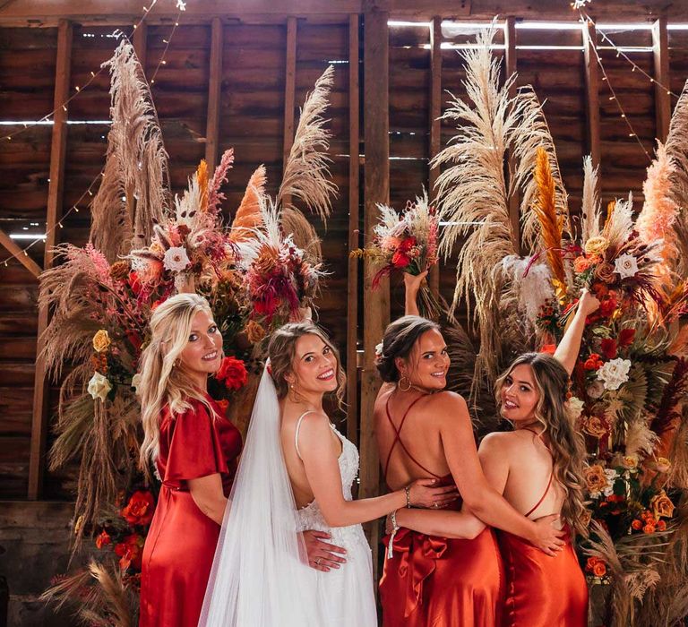 Bride in corset back wedding dress and veil stands looking over her shoulder with bridesmaids in mixed orange satin bridesmaid dresses by rustic floral installation at barn wedding ceremony in Essex