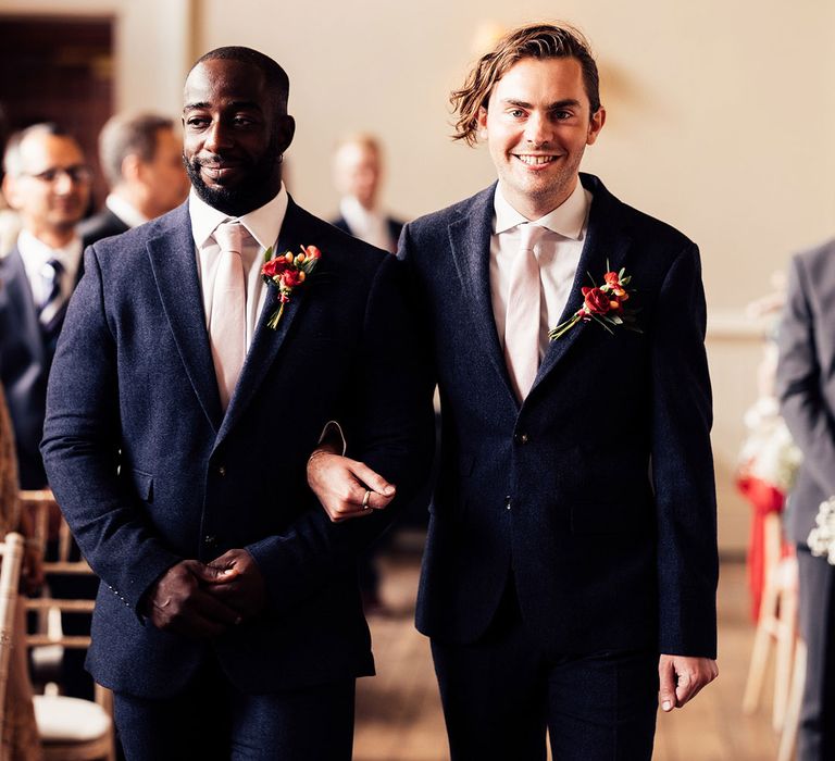 Bridesmen in blue suits, baby pink ties and floral buttonholes walk down the aisle of Elmore Court wedding ceremony room 