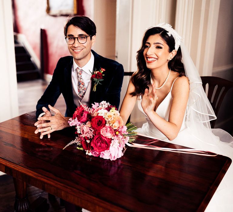 Bride in feathered headband, pearl necklace and wedding dress with plunging neckline sits with groom in dark suit and floral Gucci tie at table with red and pink bridal bouquet during wedding ceremony 