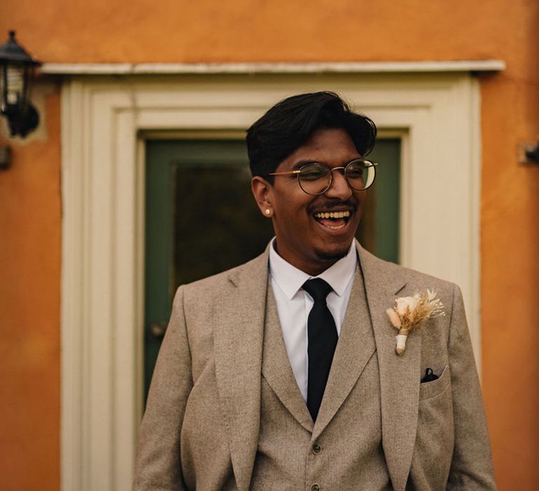 Groom wears beige three piece suit complete with black tie and dried floral buttonhole 