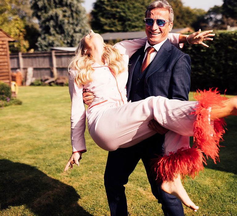 Man in blue three piece suit, orange tie and sunglasses picks up bride in pink pyjamas with red fur trim in garden