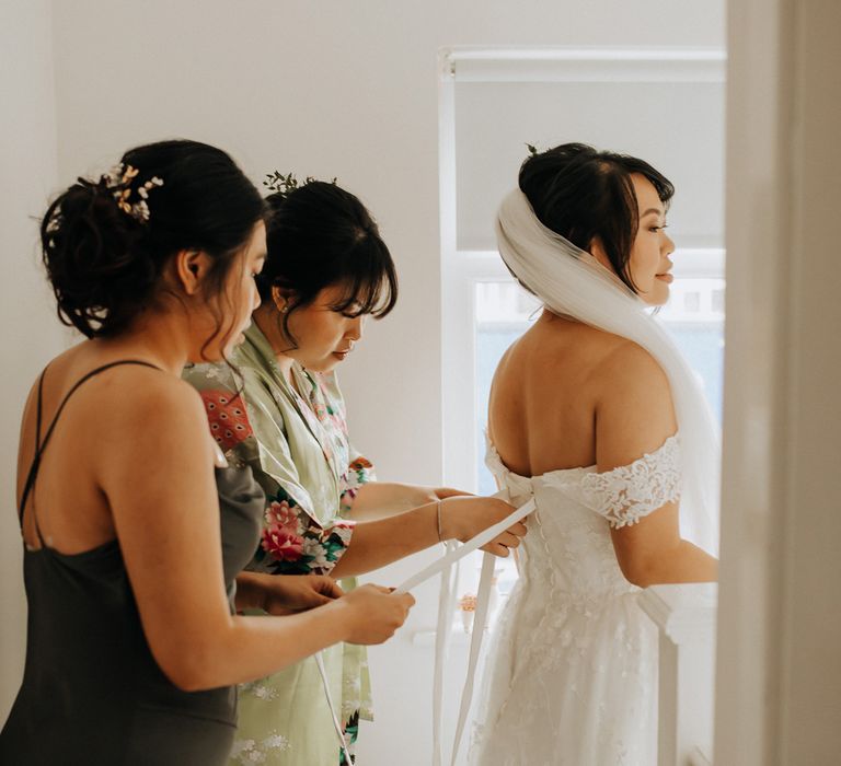 Bride in bardot corset back wedding dress and veil has the back of her dress tied by two bridesmaids before wedding