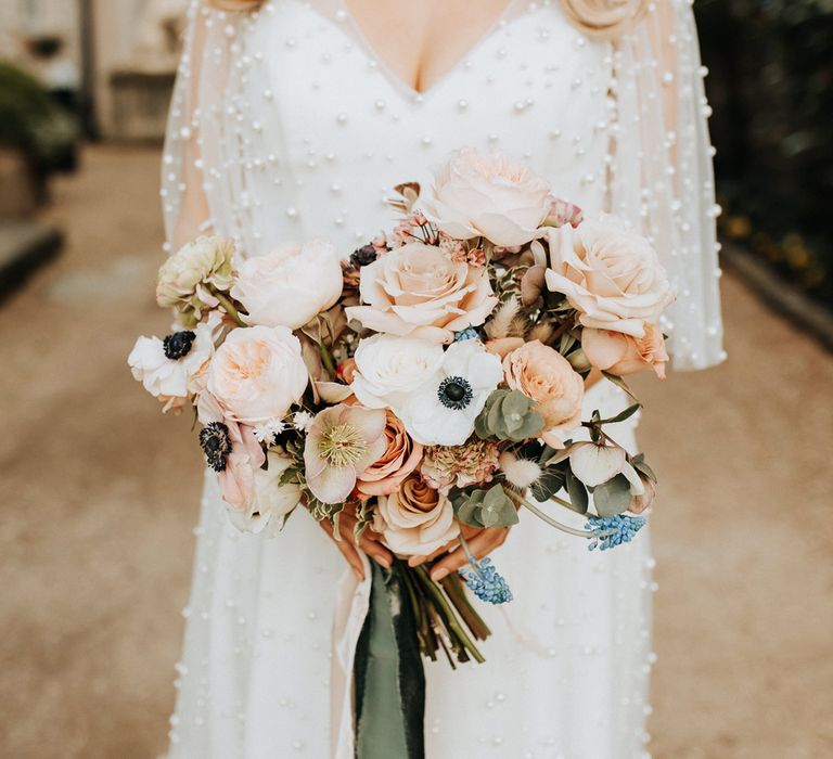 Bride in white wedding dress with pearl mesh overlay stands outside holding mixed pastel wedding bouquet with roses and anemones