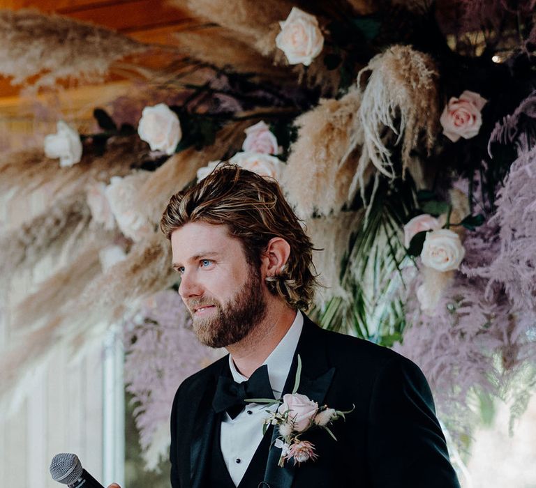 Groom in black tuxedo with a pink rose buttonhole flower giving his wedding speech 