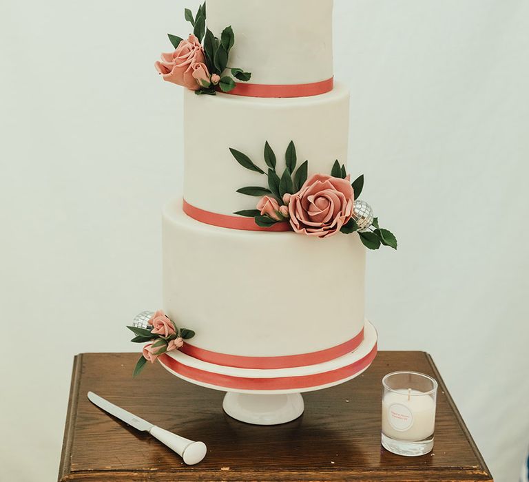 Three-tier wedding cake with white icing and pink borders with pink roses and mini disco ball decorations 