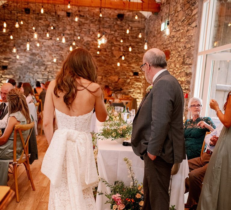 Bride in lace wedding dress with large bow sands with wedding guests at their reception