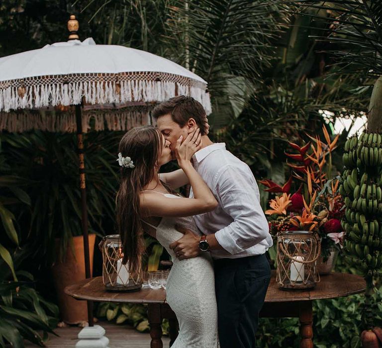 Bride in lace fitted wedding dress with fishtail train kisses groom in white shirt and black trousers