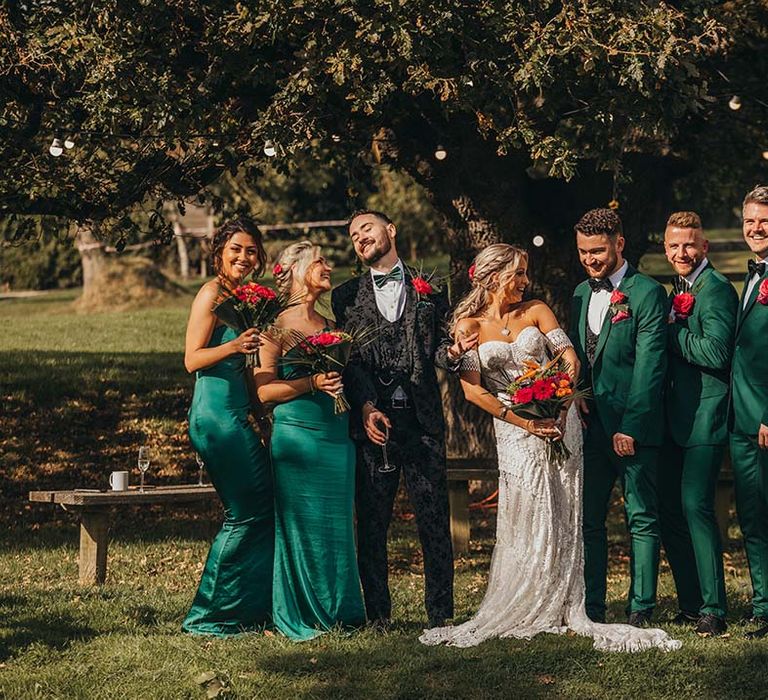 Groomsmen in emerald green black tie suits with bridesmaids in matching emerald green one shoulder dresses with bride and groom 