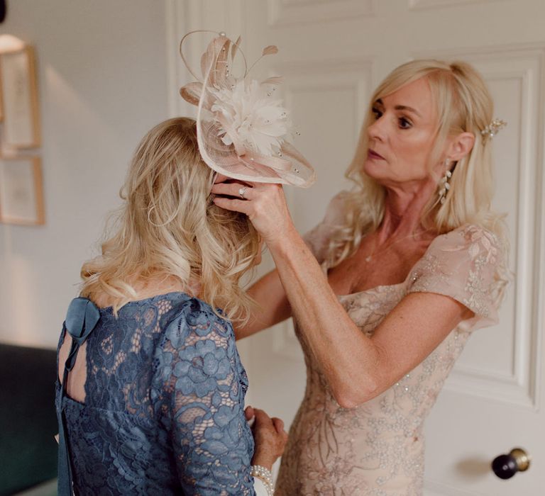 Wedding guest in sparkly gold dress places light pink fascinator on another guest's head in blue lace dress 