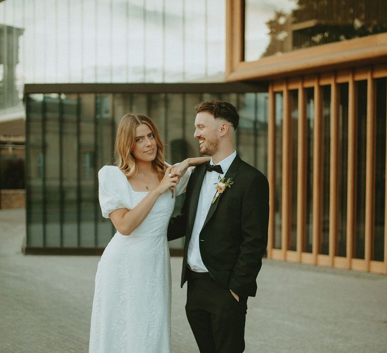 Groom in black tie smiles as he looks at the bride who rests her arm on his shoulder in puff sleeve patterned wedding dress