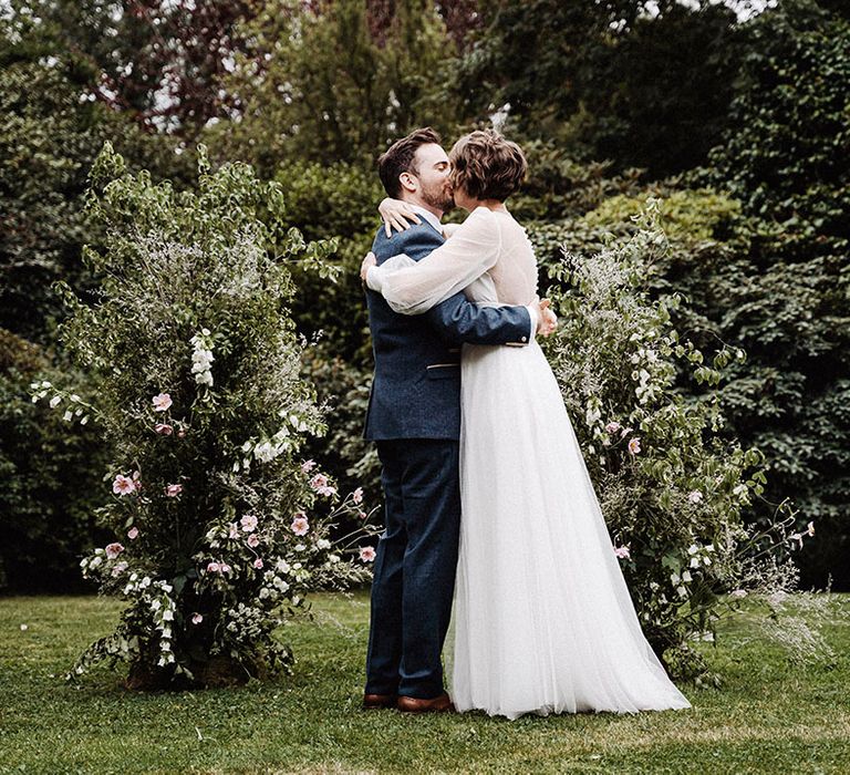 Groom in blue suit and gold patterned tie with handkerchief kisses bride in long sleeve wedding dress in front of flower columns