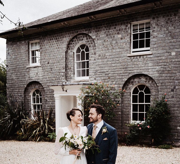 Bride in sheer long sleeve Sassi Holford wedding dress with groom in blue suit outside wedding venue, Treseren