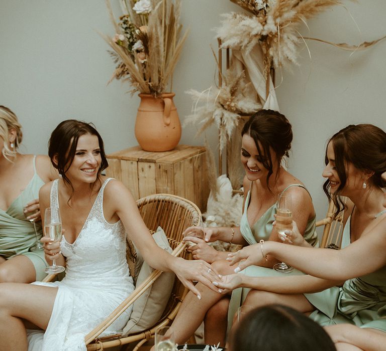 Bride shows off her silver engagement ring to her bridesmaids in sage green satin wrap dresses