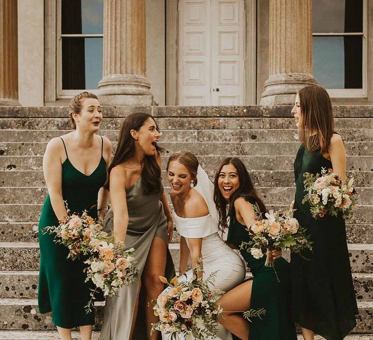 Bridesmaids in different shades of forest green bridesmaid dresses with bride in off the shoulder gown on steps of the wedding venue 