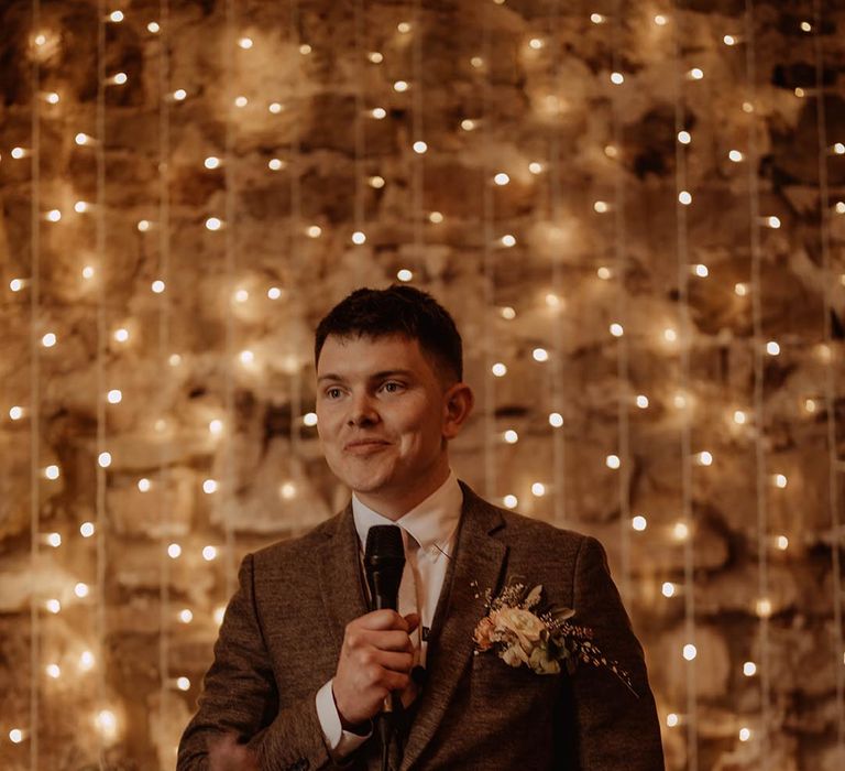 Groom reads out his speech in three piece brown suit with fairy light backdrop at Eden Barn