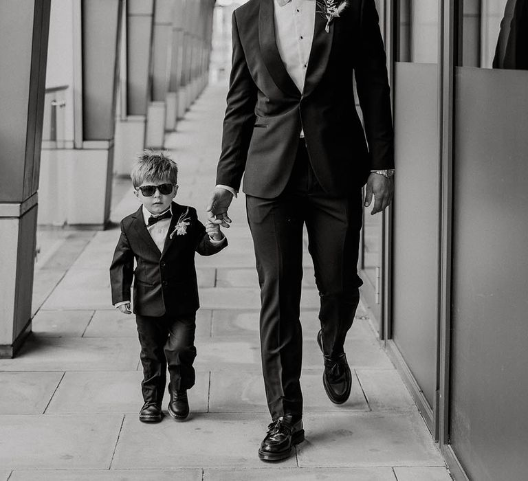 Groom in black tie with light blue bow tie walks with son in black tie and sunglasses 