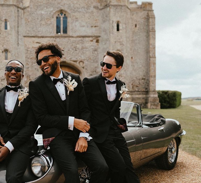 Groomsmen sitting on a wedding car in tuxedos wearing sunglasses 