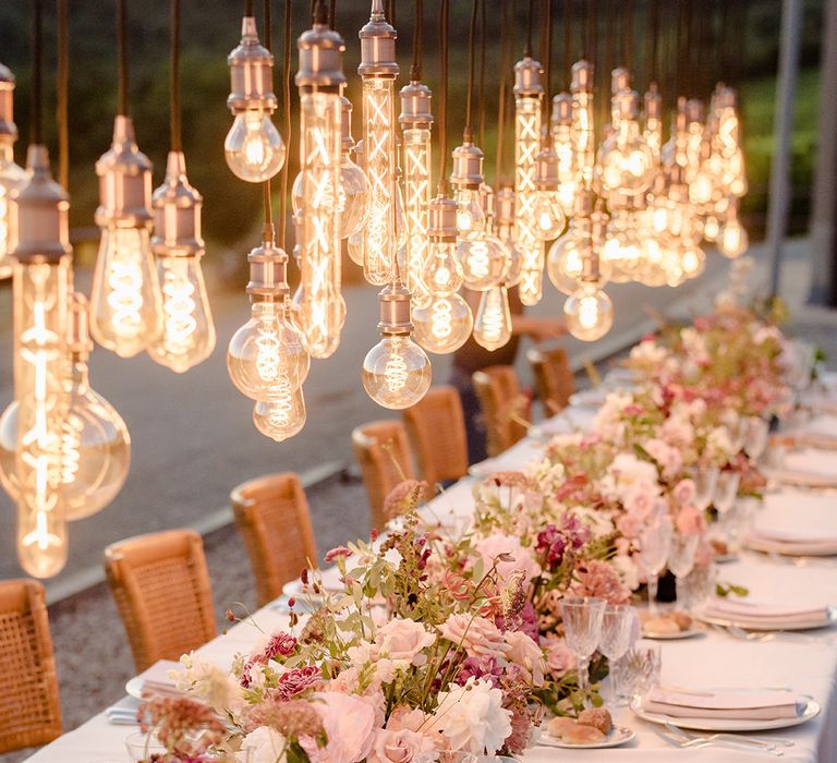 Decorative Edison bulbs installation hangs above head table complete with pastel pink and purple floral bouquets lining the middle 