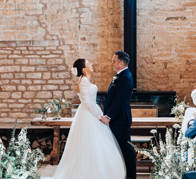 Groom holds the bride's hands as they both laugh at the altar of the Old Gore Barn with groom in a blue suit and bride in tulle ruffle wedding dress