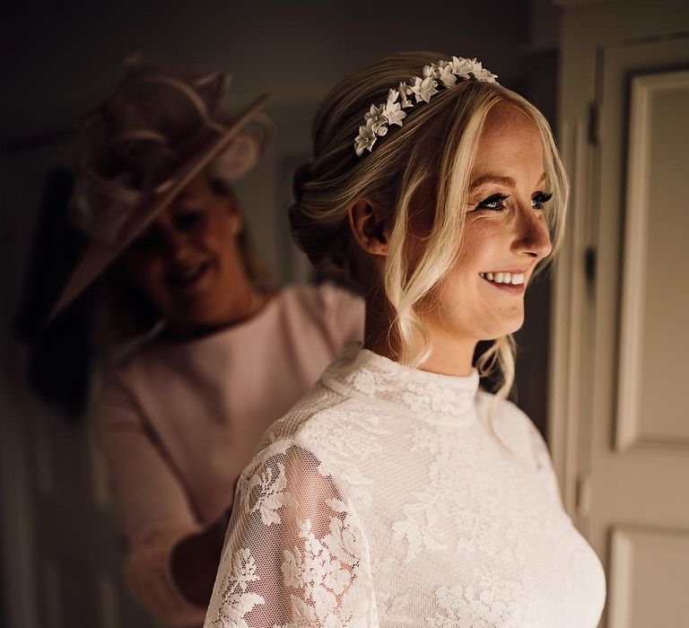 Bride in lace wedding dress with a high neck and white flower headband gets assistance from the mother of the bride in a pink outfit 