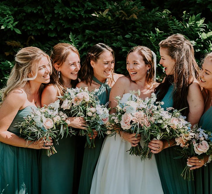 Bride stands with her bridesmaids who wear green dresses in different styles and hold pastel bouquets 