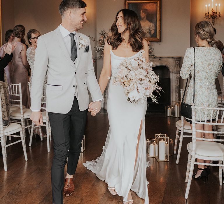 Bride carrying a white and blush pink bouquet and walking with interlocked fingers with the groom as they exit their ceremony 