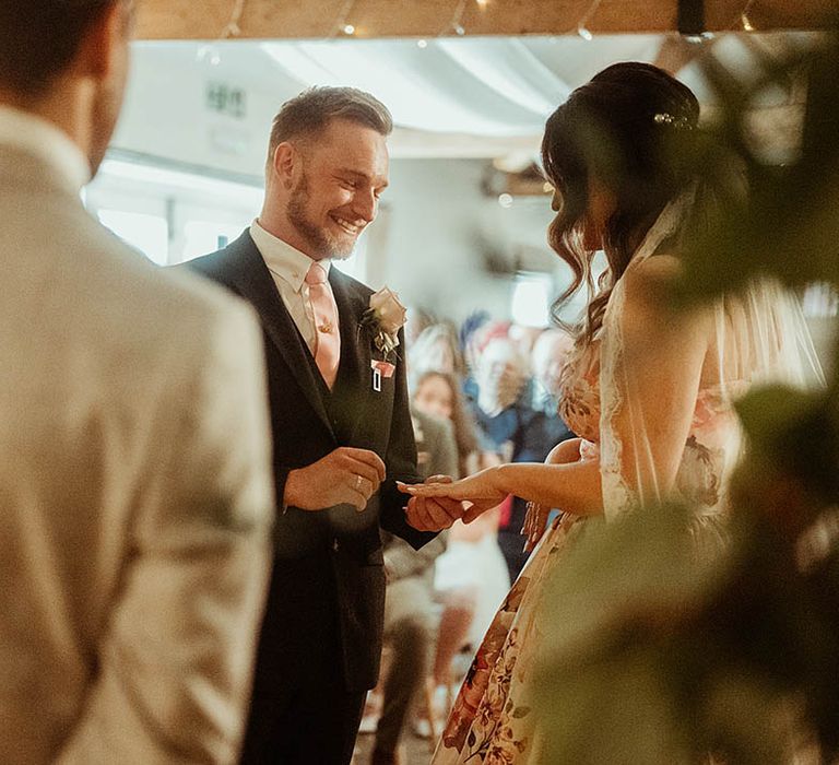 Groom wearing a suit and pink tie puts the ring on the bride's finger 