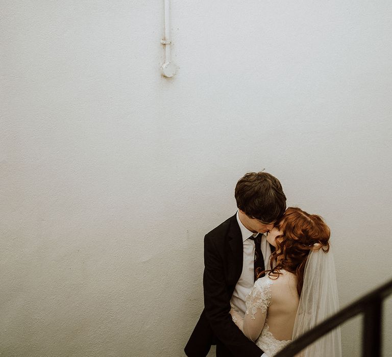 Bride wears her red hair in curls alongside floor-length veil whilst kissing her groom outdoors 