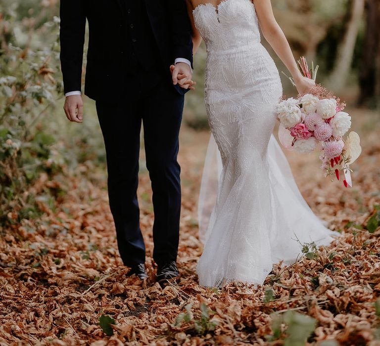 Bride & groom walk through woods during back garden wedding as bride carries white and pink floral bouquet 